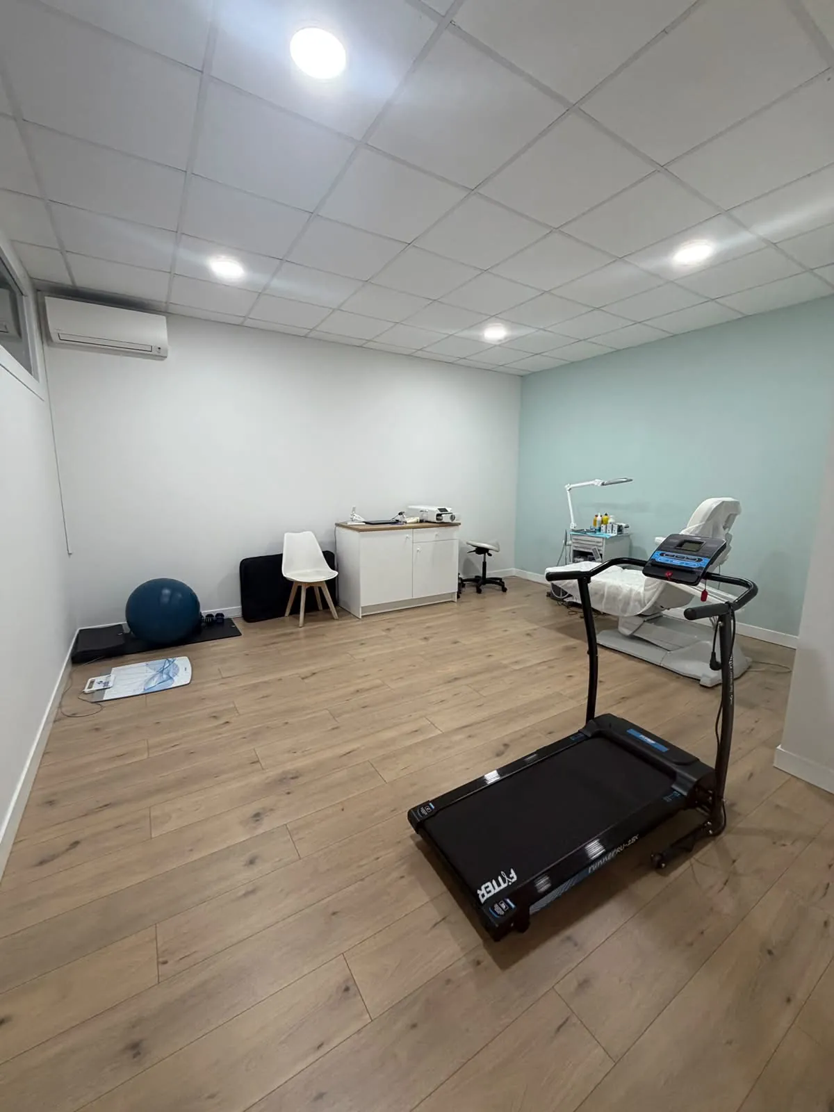 Sala de tratamiento y ejercicio terapéutico con cinta de andar, fitball, colchonetas y el sillón de podología al fondo. Clínica Paula Vega, Leganés