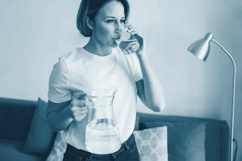 Mujer bebiendo un vaso de agua en casa con una jarra en la mano