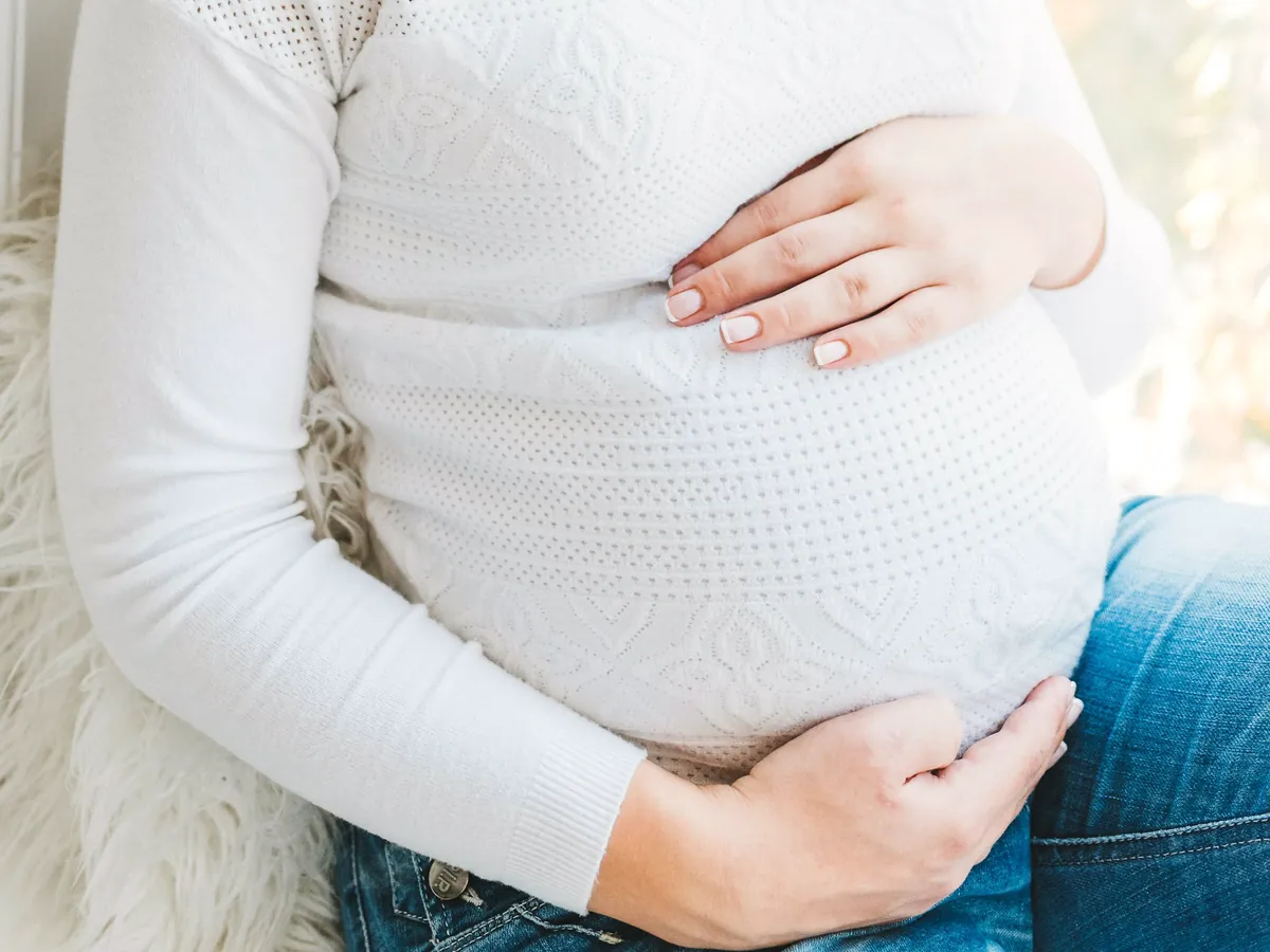 Mujer embarazada vestida de blanco con las manos apoyadas sobre la barriga, sentada junto a una ventana